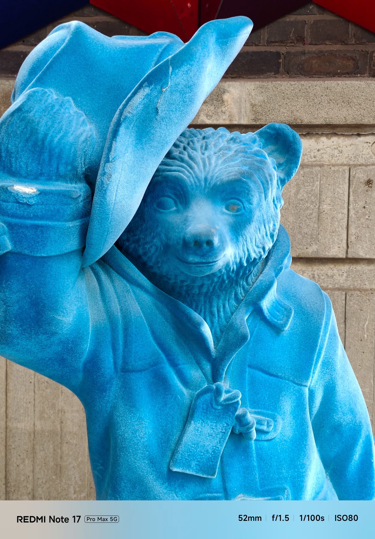 Bright blue Paddington Bear statue tipping its hat in front of a brick wall.