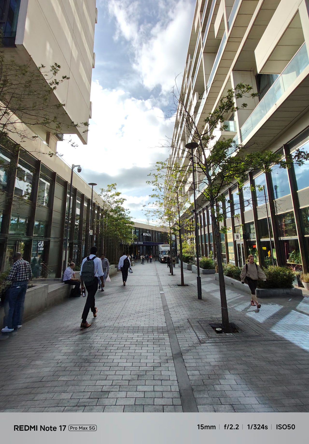 Wide-angle view along a pedestrian walkway between modern buildings under a partly cloudy sky.